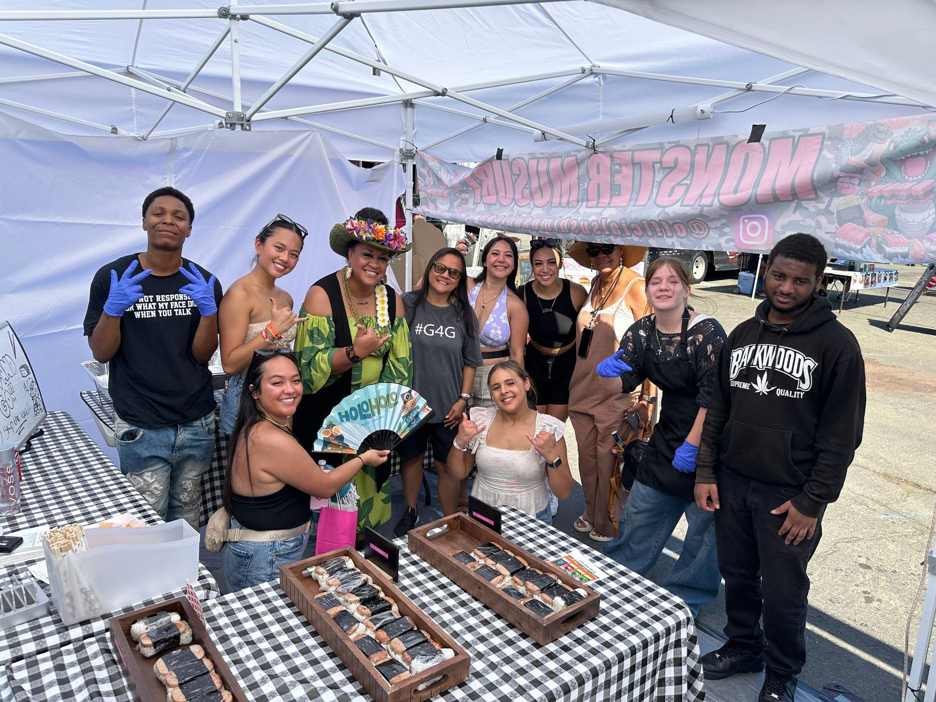 Group of people smiling under a tent at an outdoor event with food displays on checkered tables.