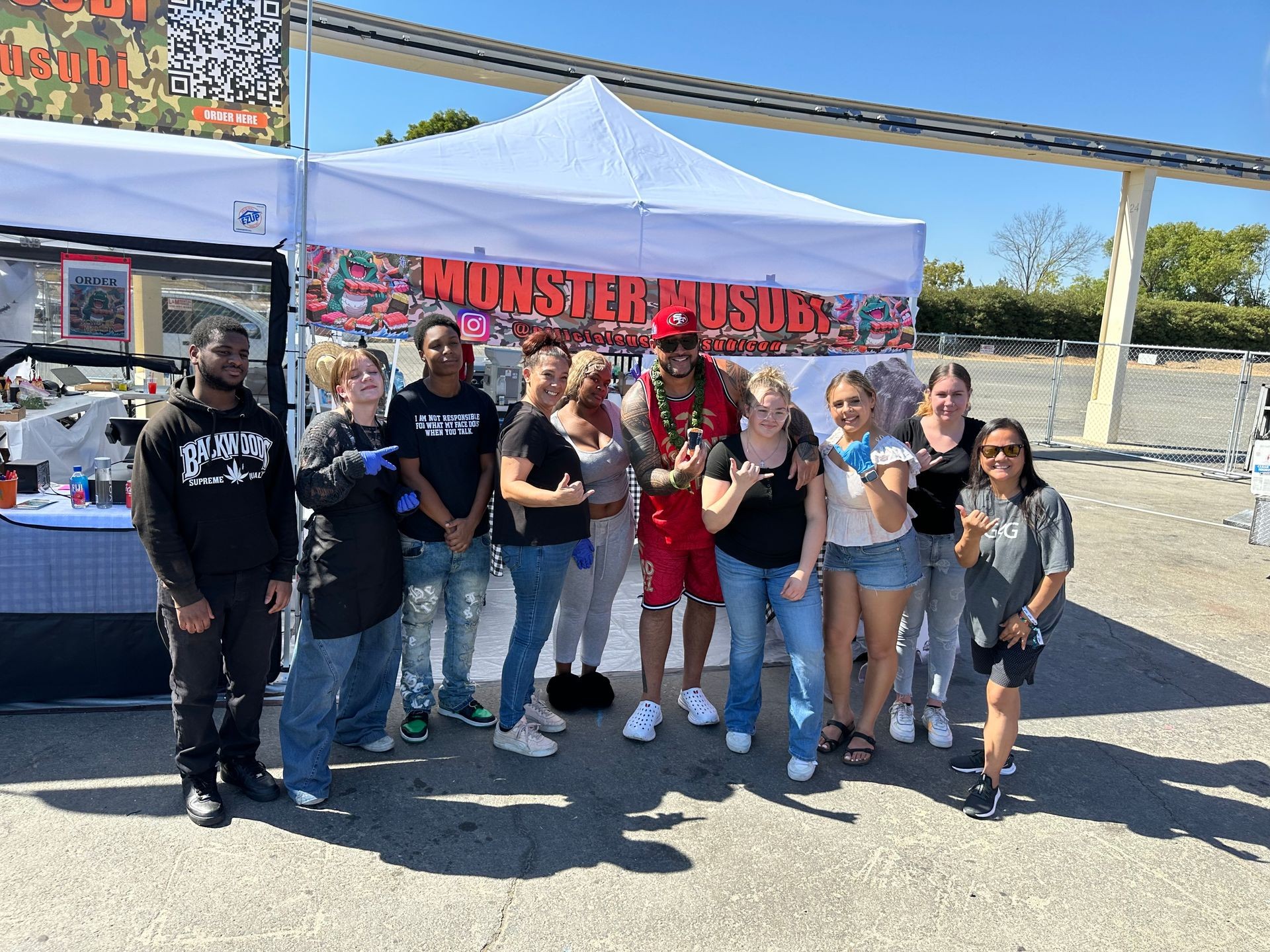 Group of people standing in front of a stall named Monster Musubi at an outdoor event.